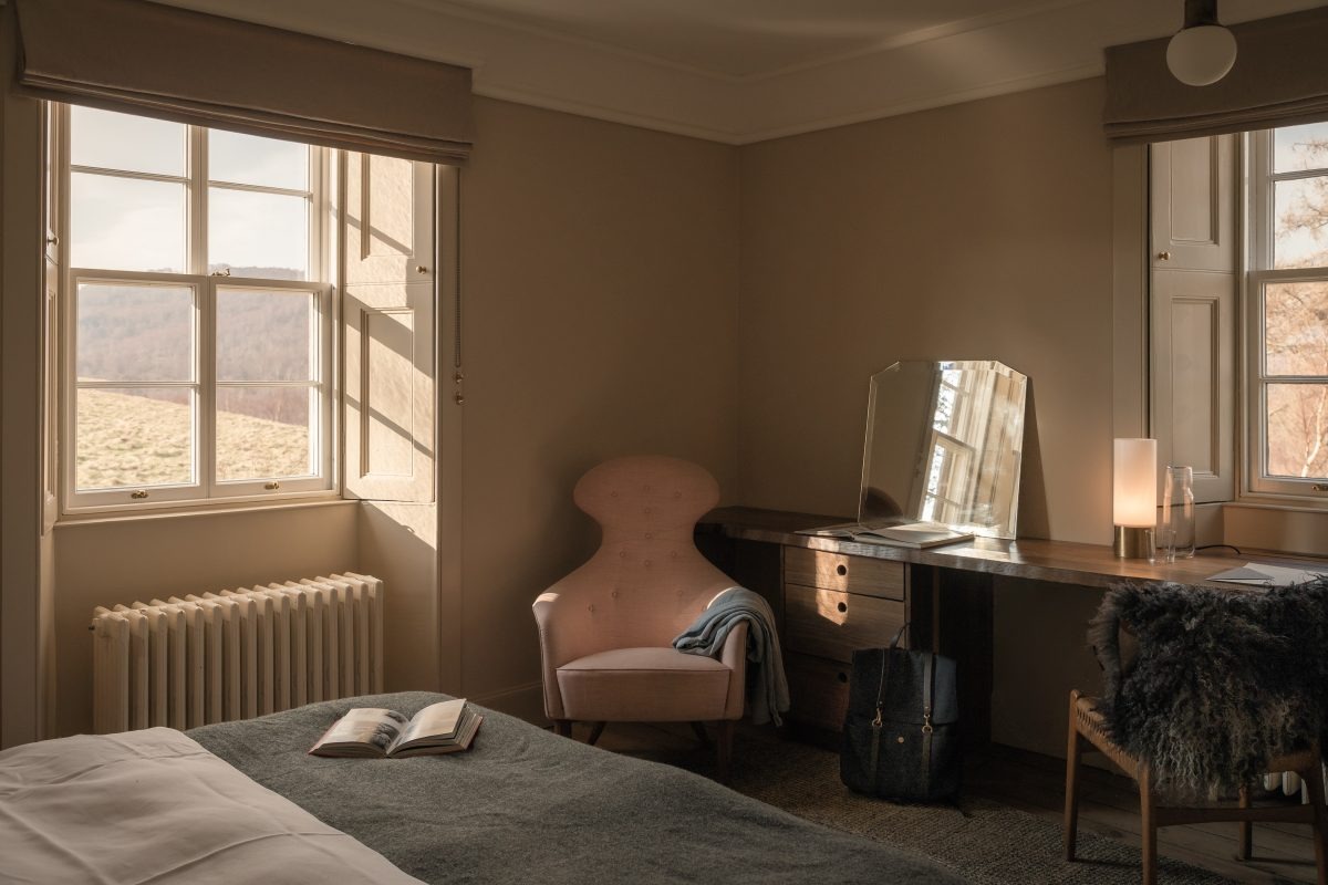 A pink armchair sits below a window looking onto hills, a dressing table with a mirror and lit lamp to the right, a bed in the foreground.