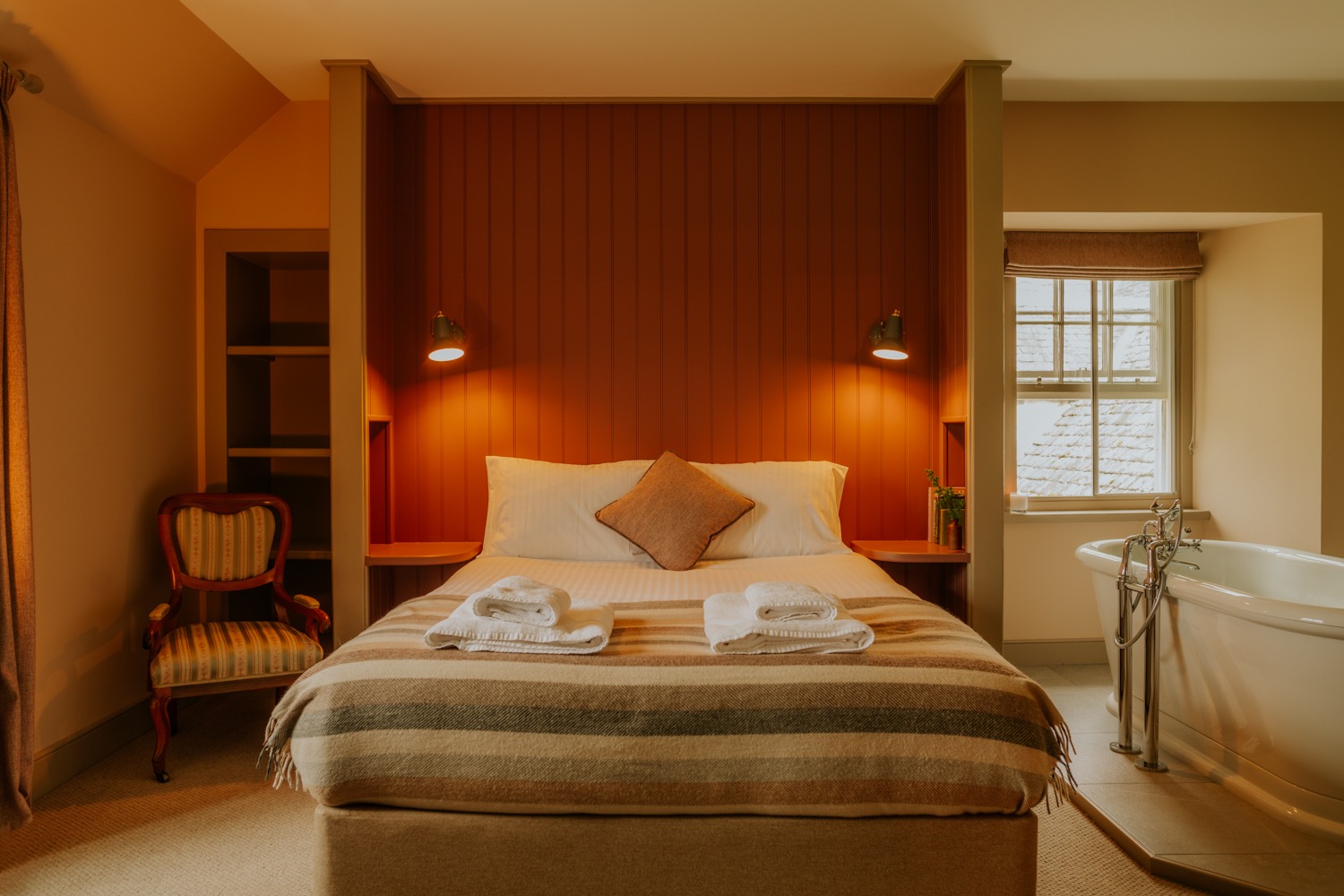 A bed with a striped throw and a cushion sits before a roll-top bath, an armchair to the left and a shuttered window to the right.