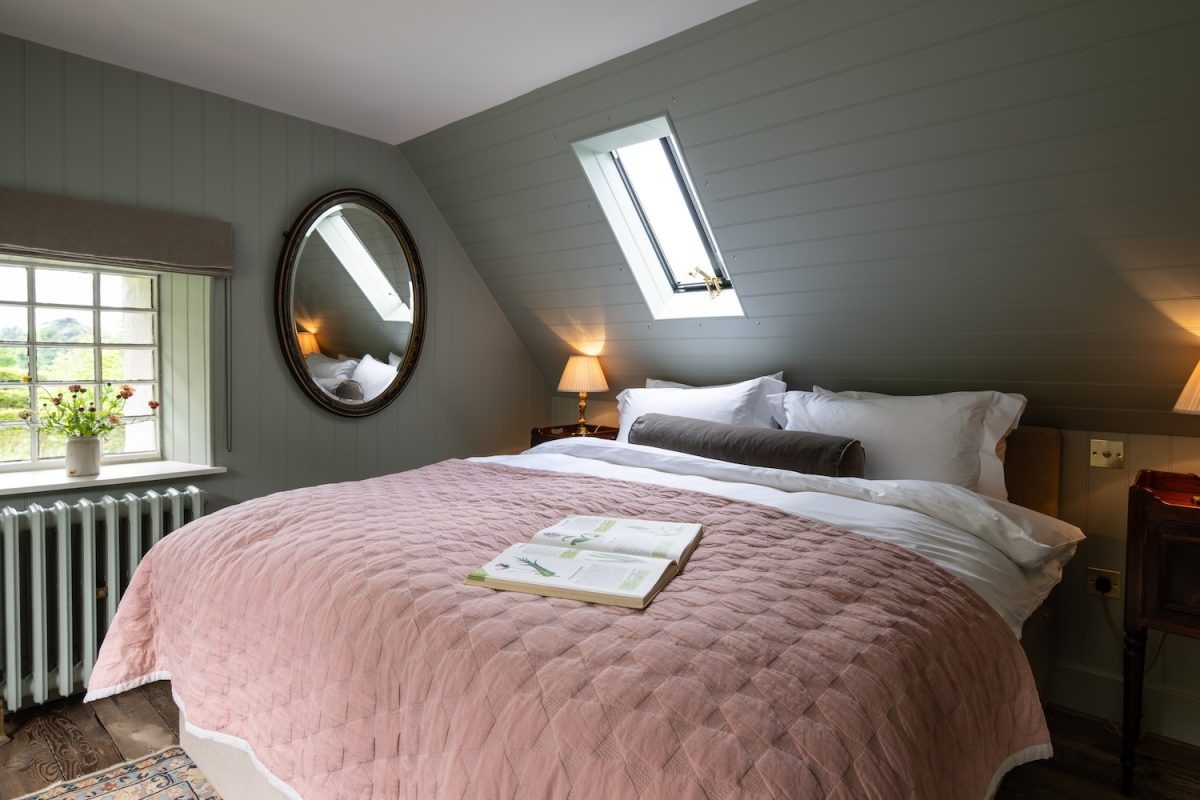 A bed dressed with a pink quilted throw and an open book sits beneath a roof window, a round mirror on the panelled wall behind.