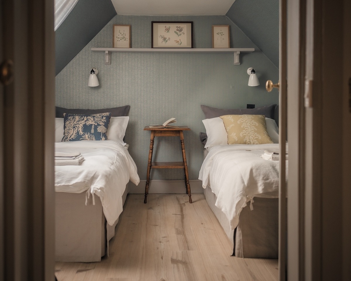 Twin beds with grey headboards flank a wooden side table beneath a shelf of framed botanical prints, on pale green striped wallpaper.