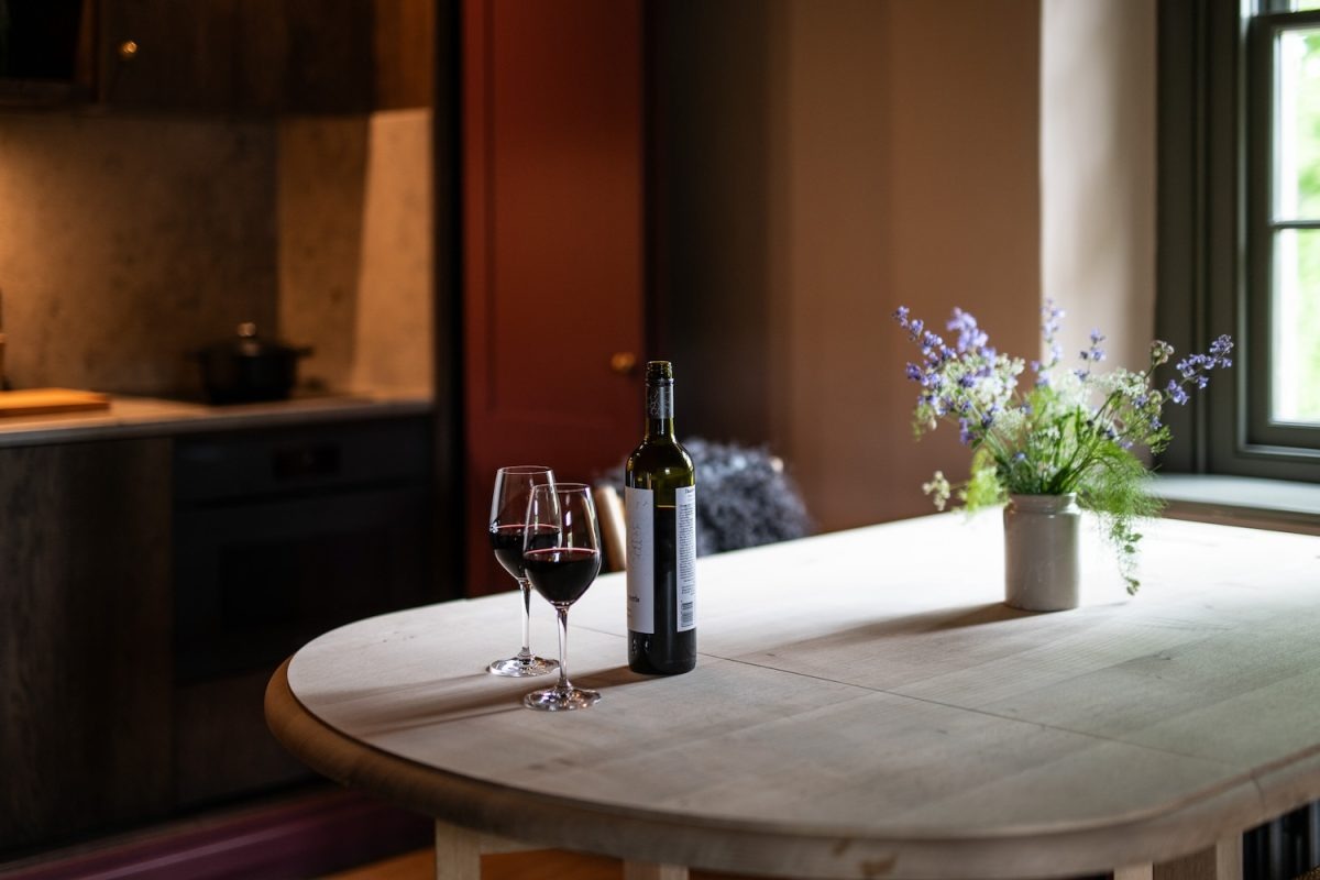 A bottle of red wine and two glasses stand on a round wooden table beside a jug of wildflowers, a kitchen visible behind.