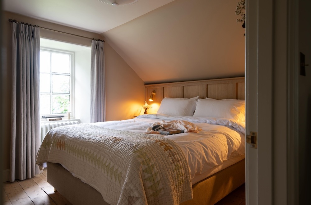 A bed with a quilted throw sits beneath a panelled headboard, a lit lamp on the bedside table and curtains framing the window.