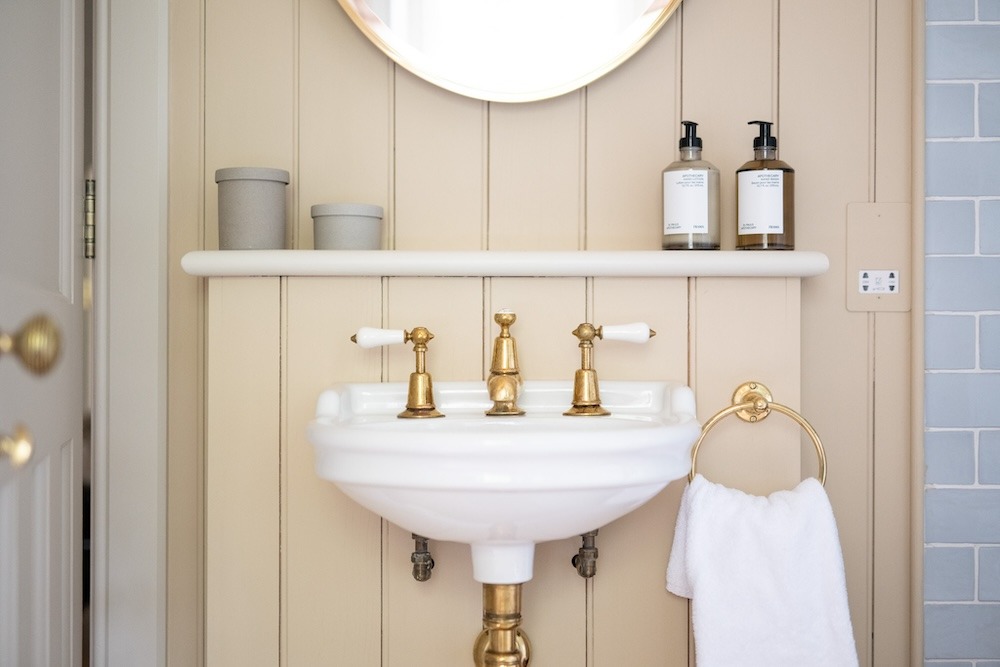 A pedestal basin with brass taps stands below a round mirror, two amber pump bottles and a folded towel on a brass ring beside it.