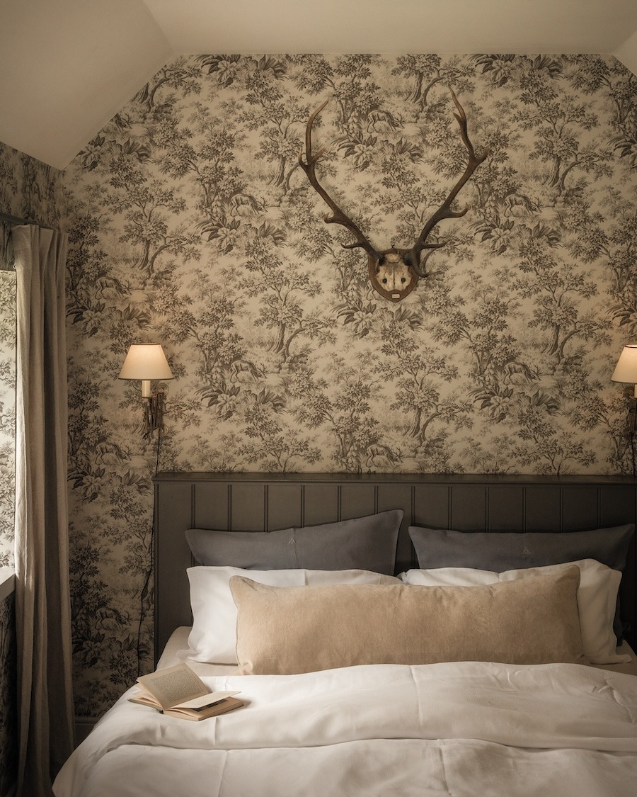 A bed with grey and white linen sits below mounted antlers on floral wallpaper, wall lamps lit either side.