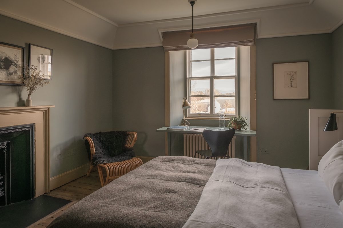 A bed with a grey throw sits before a fireplace and armchair, a writing desk beneath a window looking onto hills.