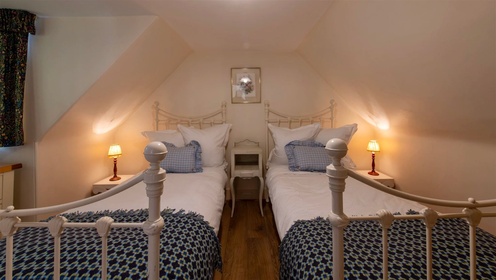 Twin white metal beds with navy patterned covers sit either side of a shared bedside table under a sloped ceiling.