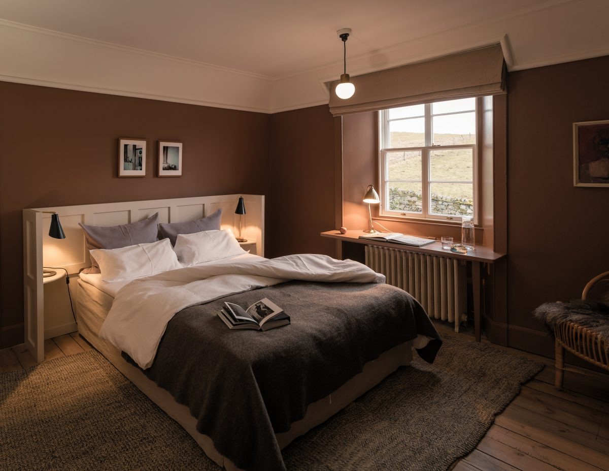 A bed with white linen and a grey throw sits between two lit wall lamps, a writing desk beneath a window to the right.