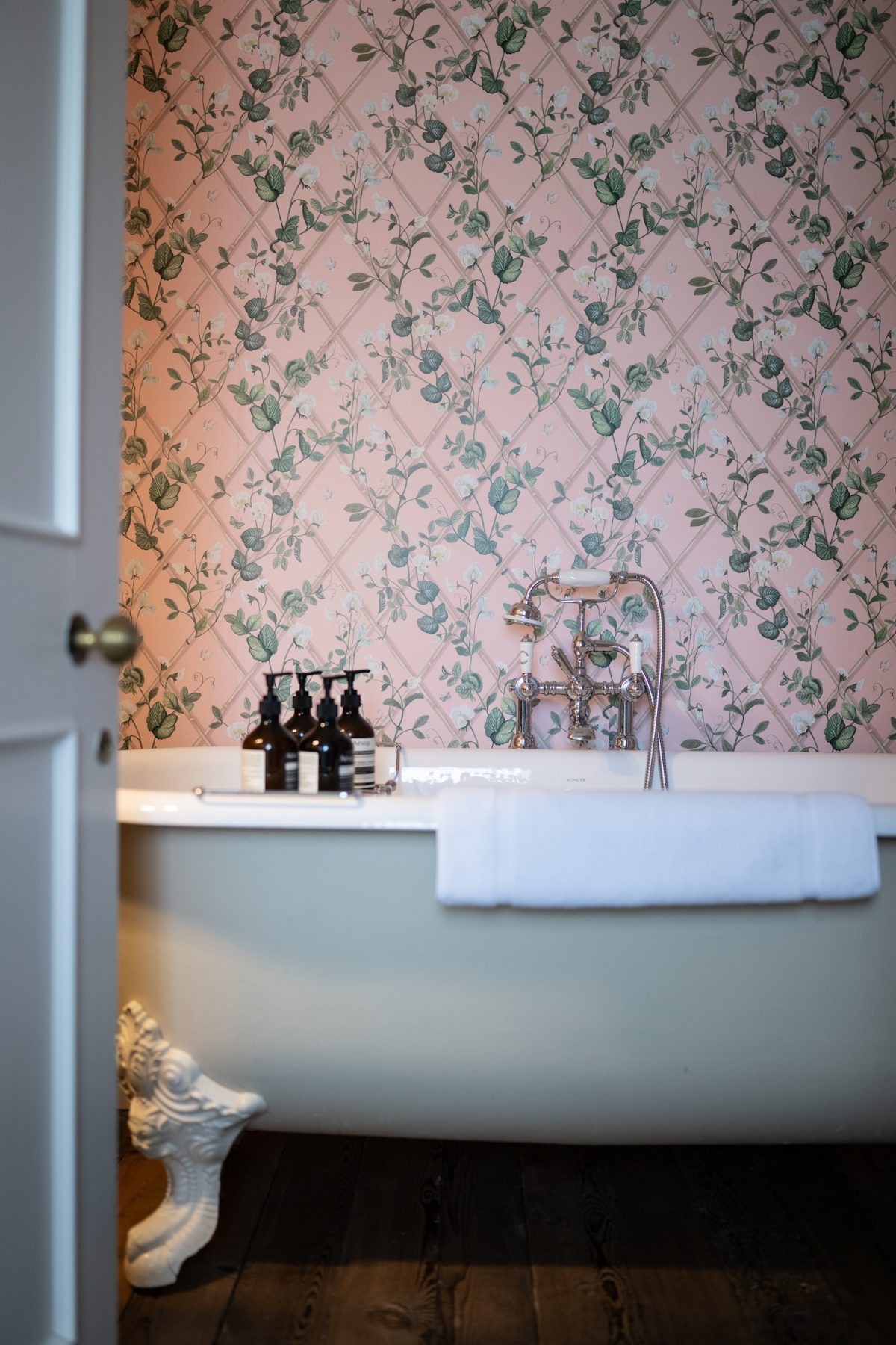 A roll-top bath stands before pink wallpaper patterned with green trellis and flowers, toiletries and a folded towel on its rim.