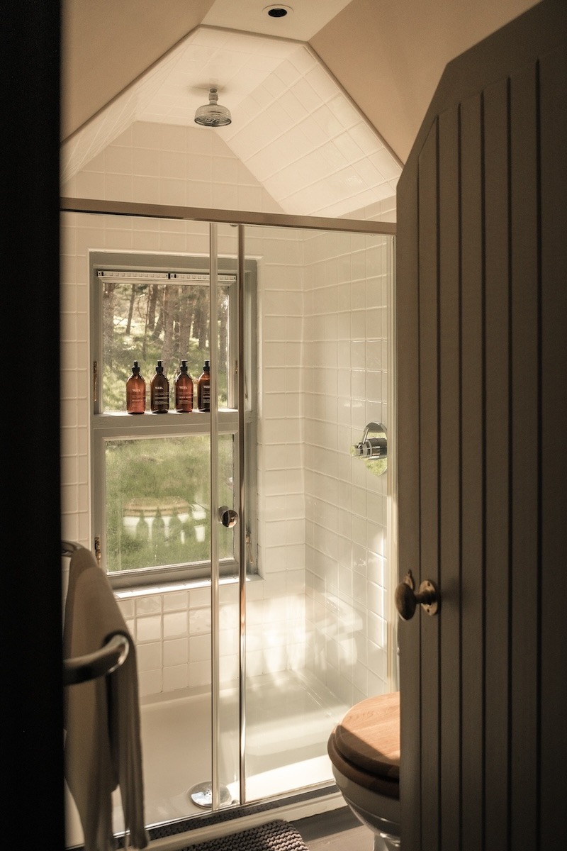 A glass shower cubicle stands beside a toilet in a white tiled bathroom, amber toiletry bottles lined on the windowsill.
