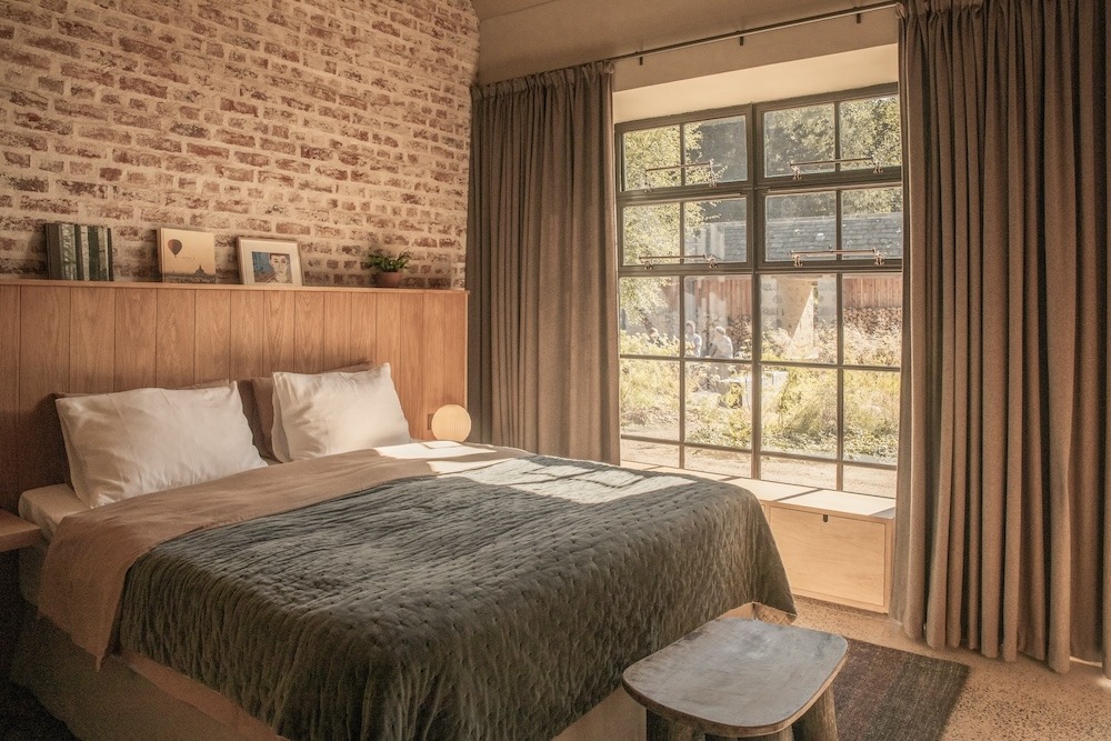 A bed with a green throw sits before an exposed brick wall and steel-framed window looking onto a garden, a wooden stool at its foot.