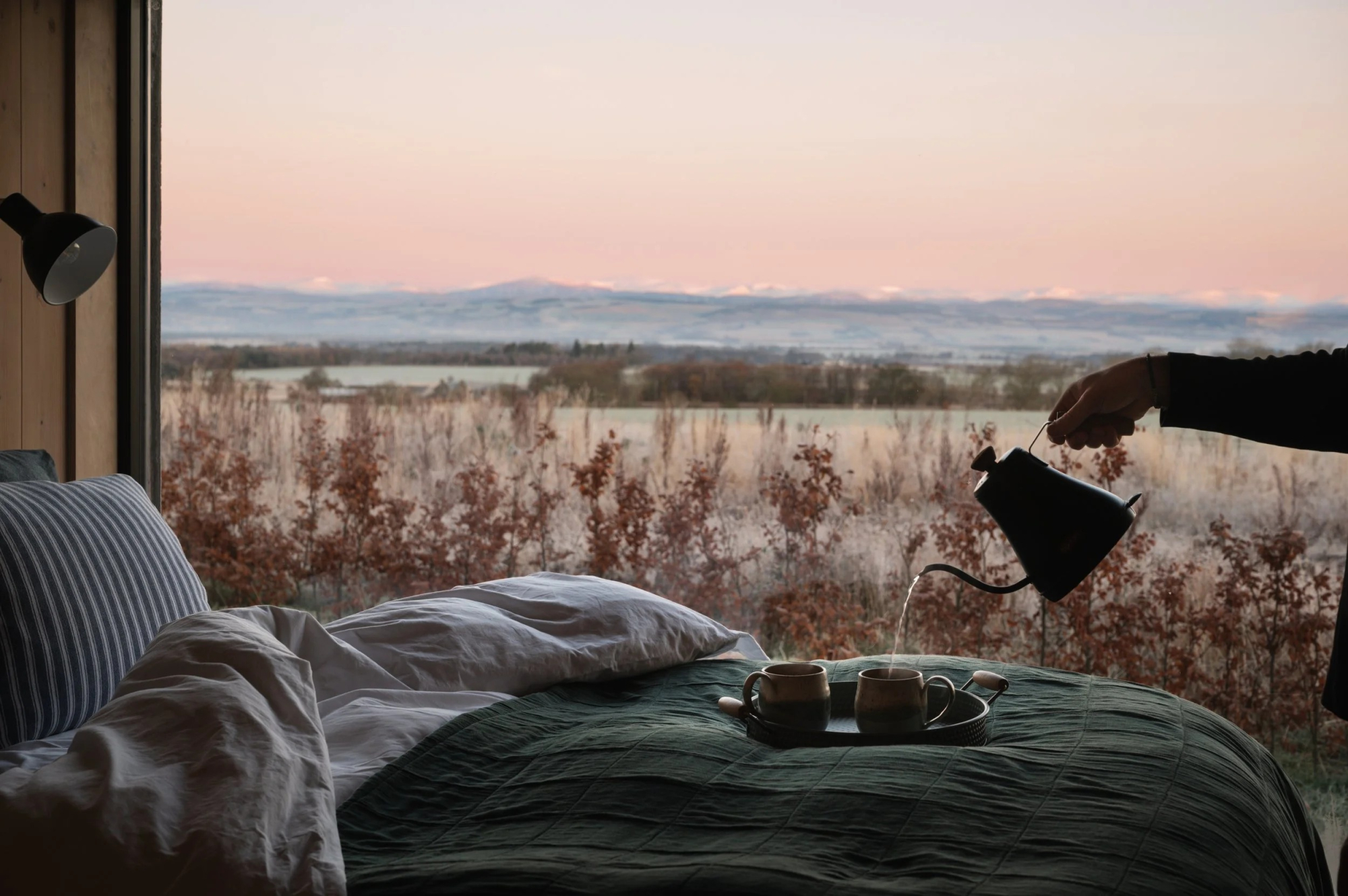 A hand pours from a black gooseneck kettle into two mugs on a bed, frosted grassland and hills visible through the window at dawn.
