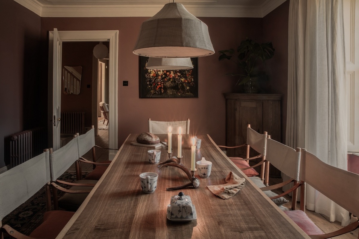 A dining table is set with candles and a small dish beneath a framed painting, director's chairs around it and a doorway leading to the hall beyond.