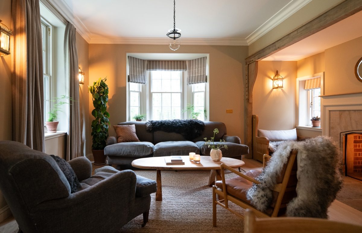 A grey corner sofa sits before a bay window dressed with striped blinds, potted plants and a leather chair with a sheepskin throw completing the room.