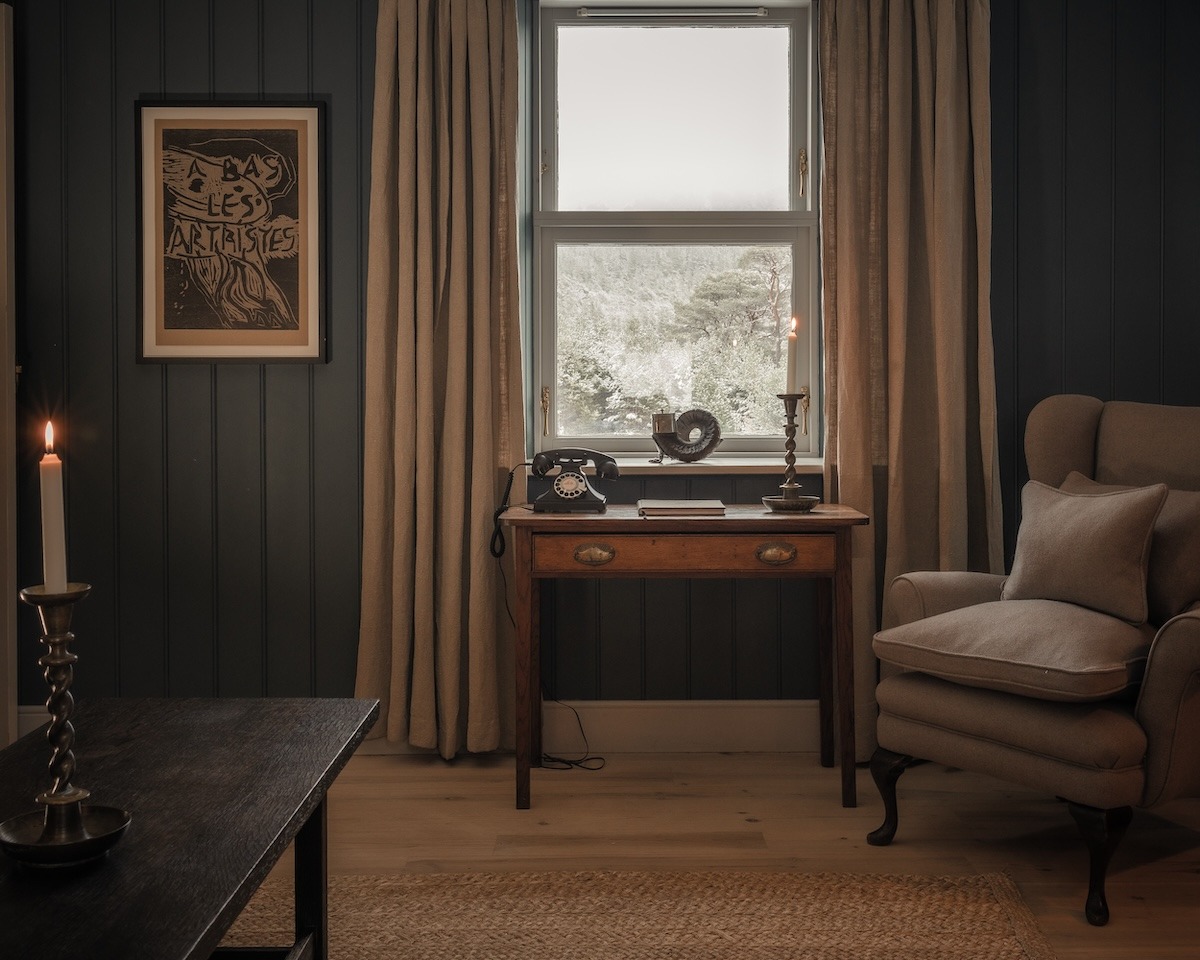 A vintage "Bal des Artistes" print hangs on a dark panelled wall above a writing desk with an antique telephone, an armchair to the right.