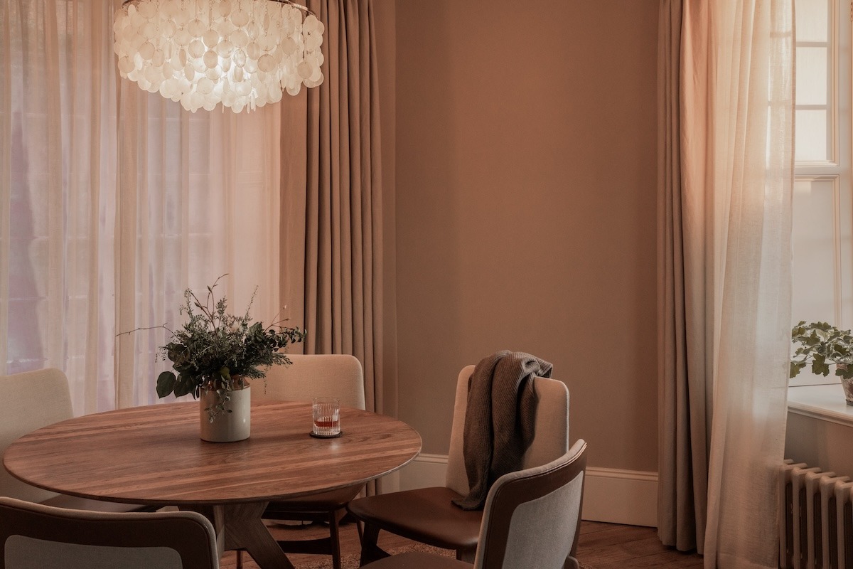 A scalloped shell chandelier hangs above a dining table set with a vase of foliage, director's chairs and curtained windows around it.