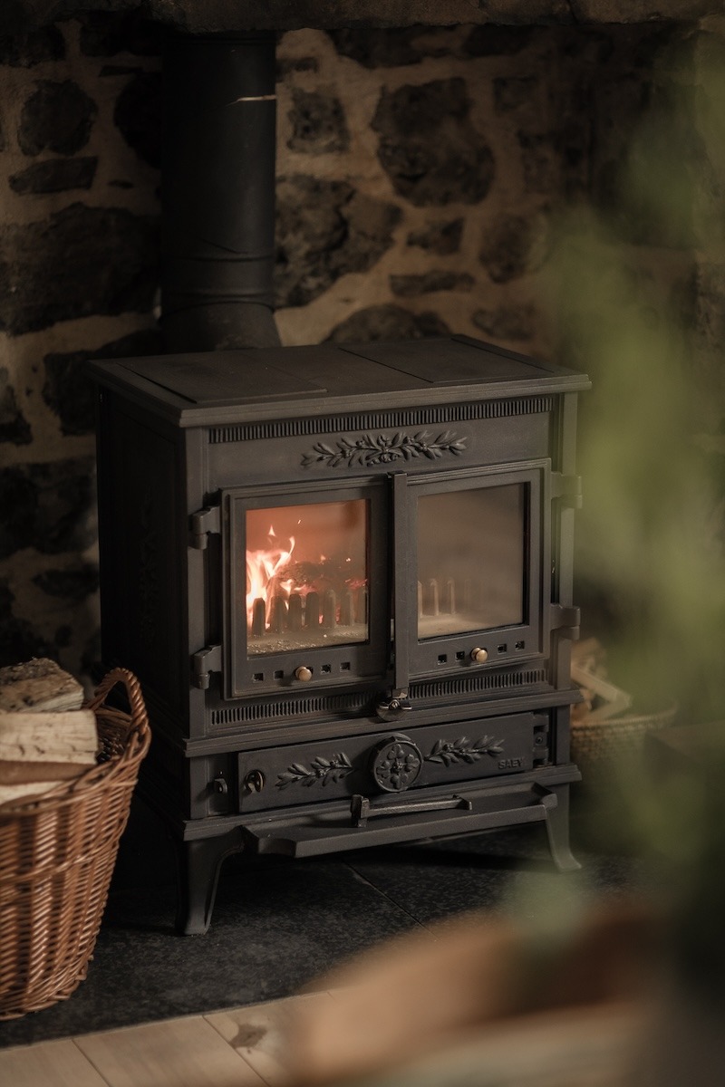 A wood burning stove is alight beneath a black flue, a wicker log basket to the side against a stone wall.
