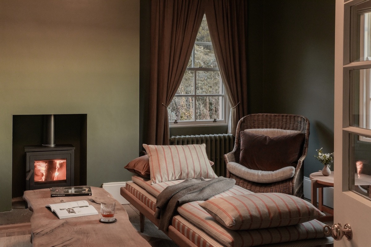 A wood-burning stove is lit beside a striped daybed and a wicker chair, a small table holding a drink and an open book.