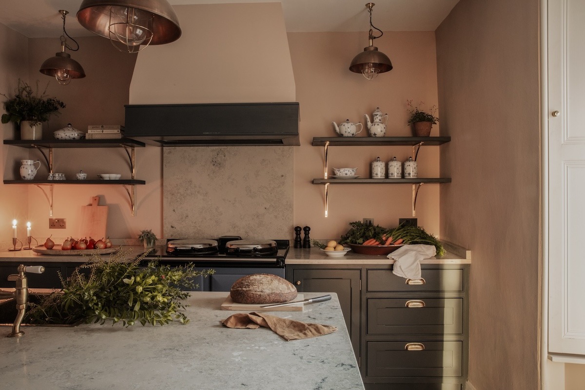 Copper pendant lights hang above a marble-topped kitchen island with dark green cabinetry, open shelving of crockery and a loaf of bread on the counter.