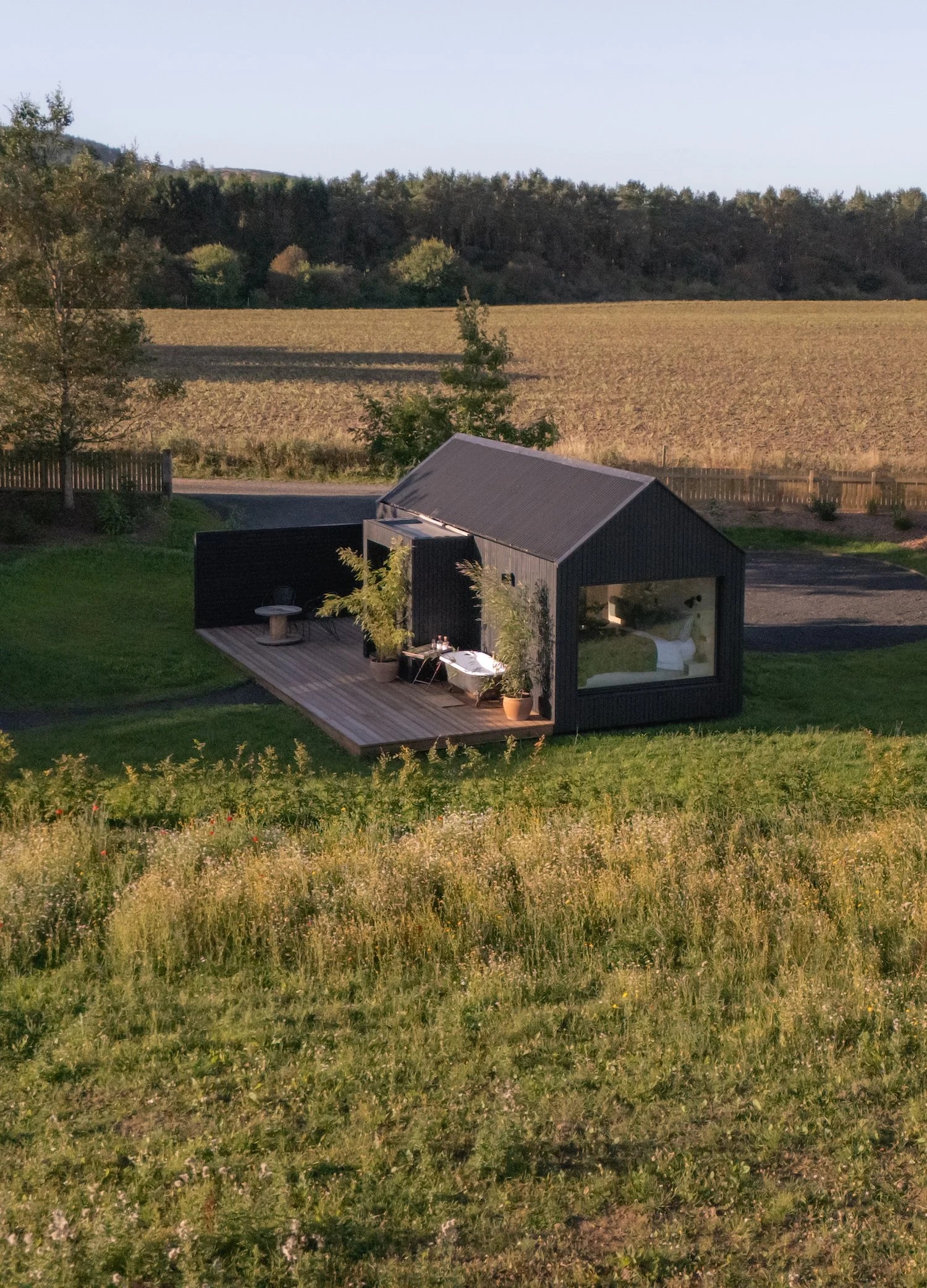 An aerial view shows a black timber cabin with a pitched roof set on a deck in an open field, a bath and seating area visible outside.