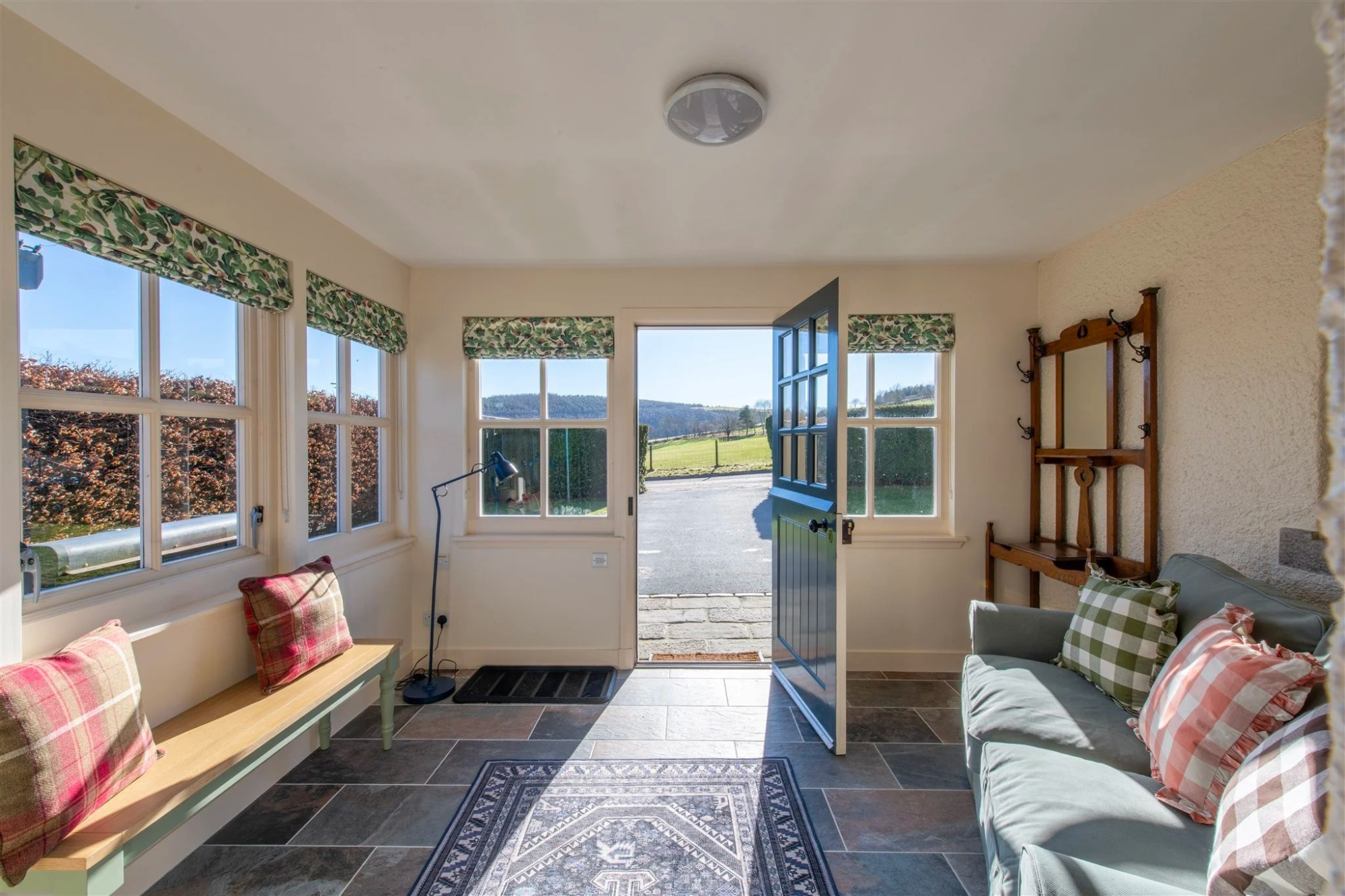 A garden room with patterned blinds and a coat rack, its stable door closed, gingham cushions on the bench and sofa.