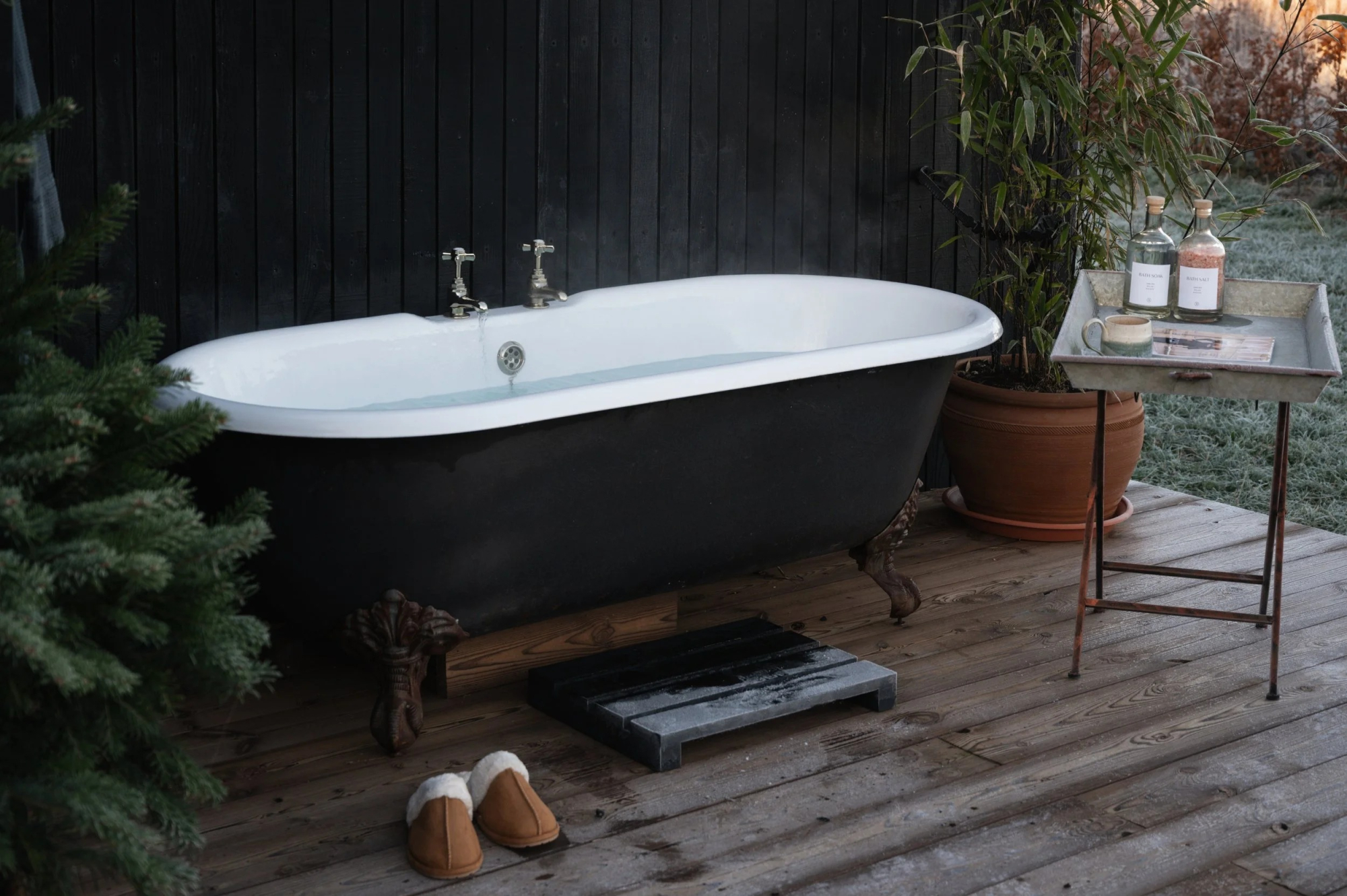 A black roll-top bath stands on a timber deck against a dark clad wall, slippers and a side table of bottles beside it.
