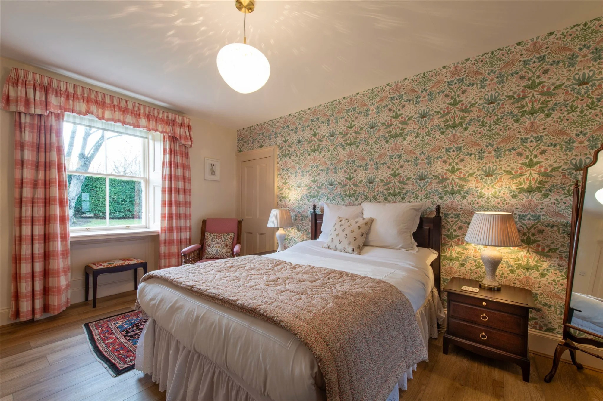 A bedroom with green floral wallpaper on one wall, a pink armchair by the window and a bed dressed in cream throws.