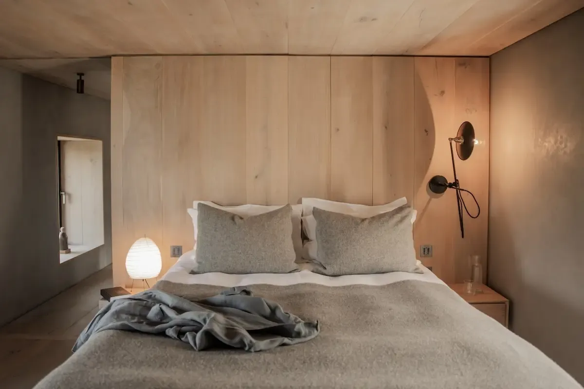 A bed with grey linen sits against timber panelling, a wall-mounted reading lamp and small round lamp on the bedside table.