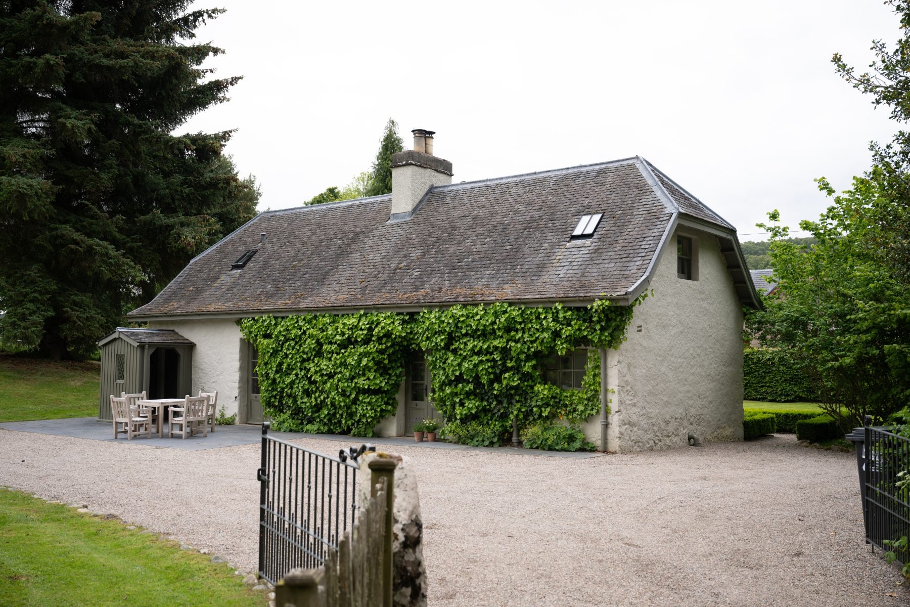 A stone cottage with a slated roof and dormer windows sits behind a gravel drive, ivy covering one gable end.