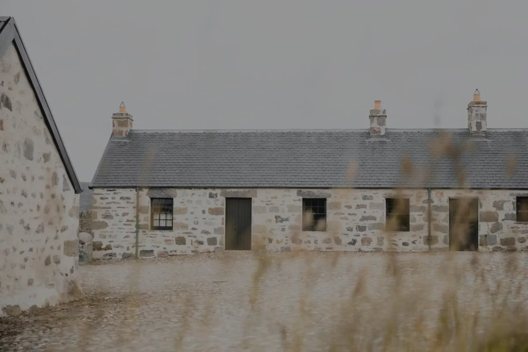 A row of stone cottages with a slated roof and chimneys stands behind a gravel yard, seen through tall grass.