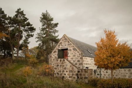 A stone barn with a red door and a steep slate roof stands among trees turning gold in autumn.