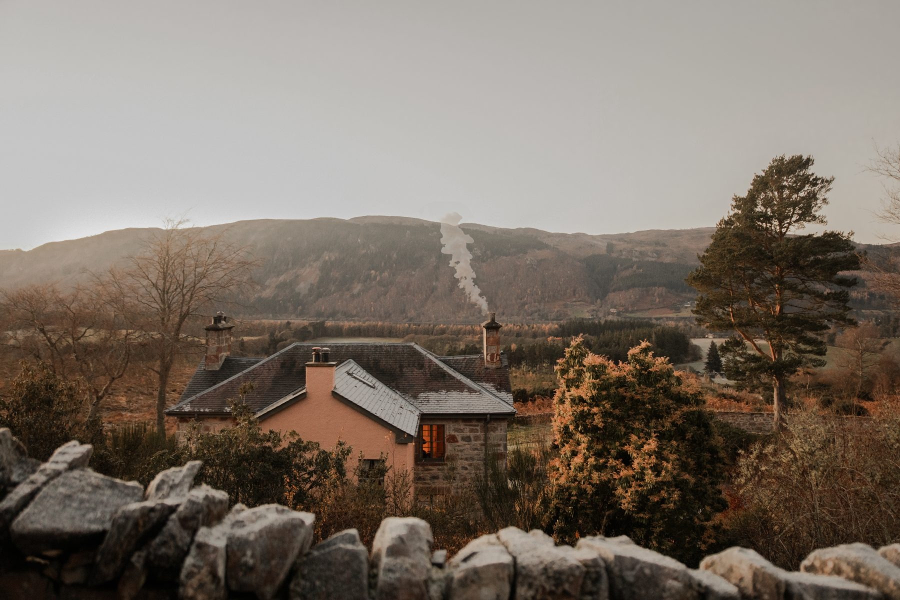 Smoke rises from the chimney of a pink-harled house with a slate roof, seen over a drystone wall amid trees and misty hills.
