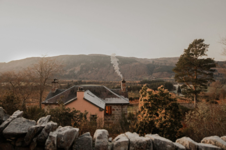 Smoke rises from the chimney of a pink-harled house with a slate roof, seen over a drystone wall amid trees and misty hills.