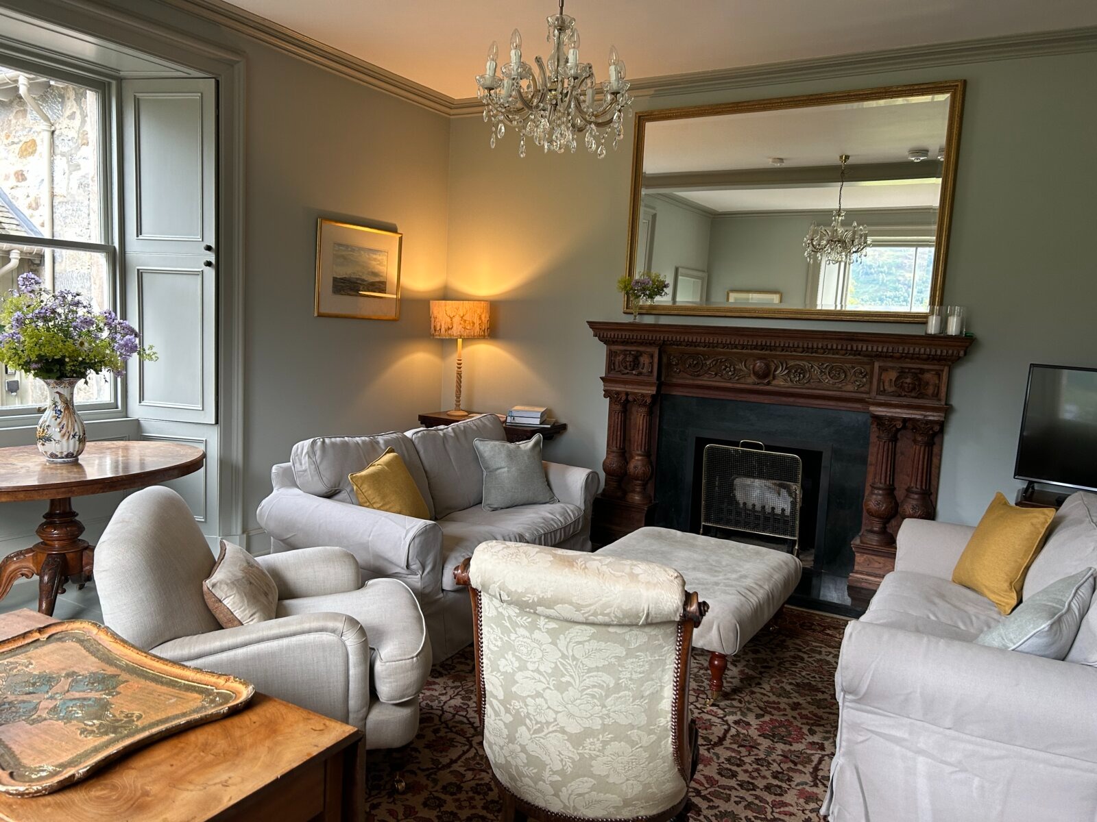 Cream sofas and armchairs surround a low table before a carved wooden fireplace with a gilt mirror above, a chandelier lit overhead.