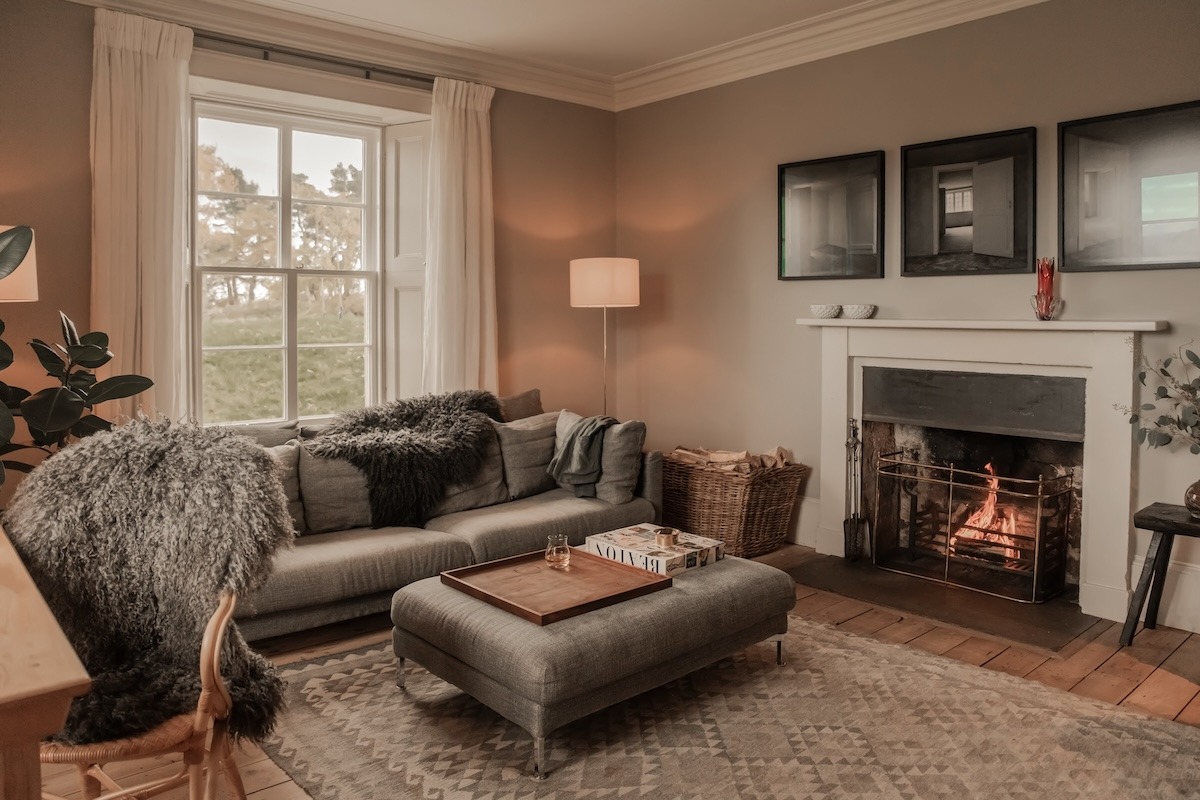 A grey sofa with a faux fur throw sits before a lit fireplace, three framed prints above the mantel and a woven basket of logs to the side.