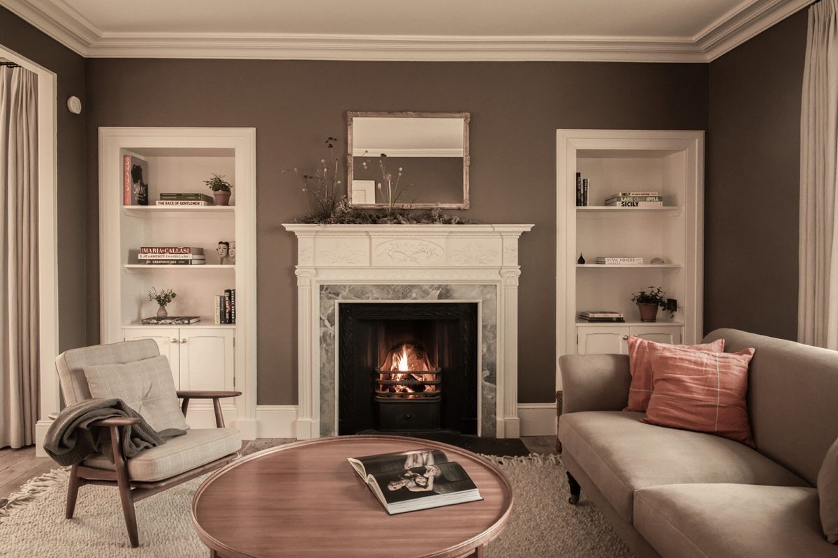 A white fireplace with a mirror above sits between built-in bookshelves, a beige sofa with a pink cushion and a wicker armchair arranged around a round coffee table.