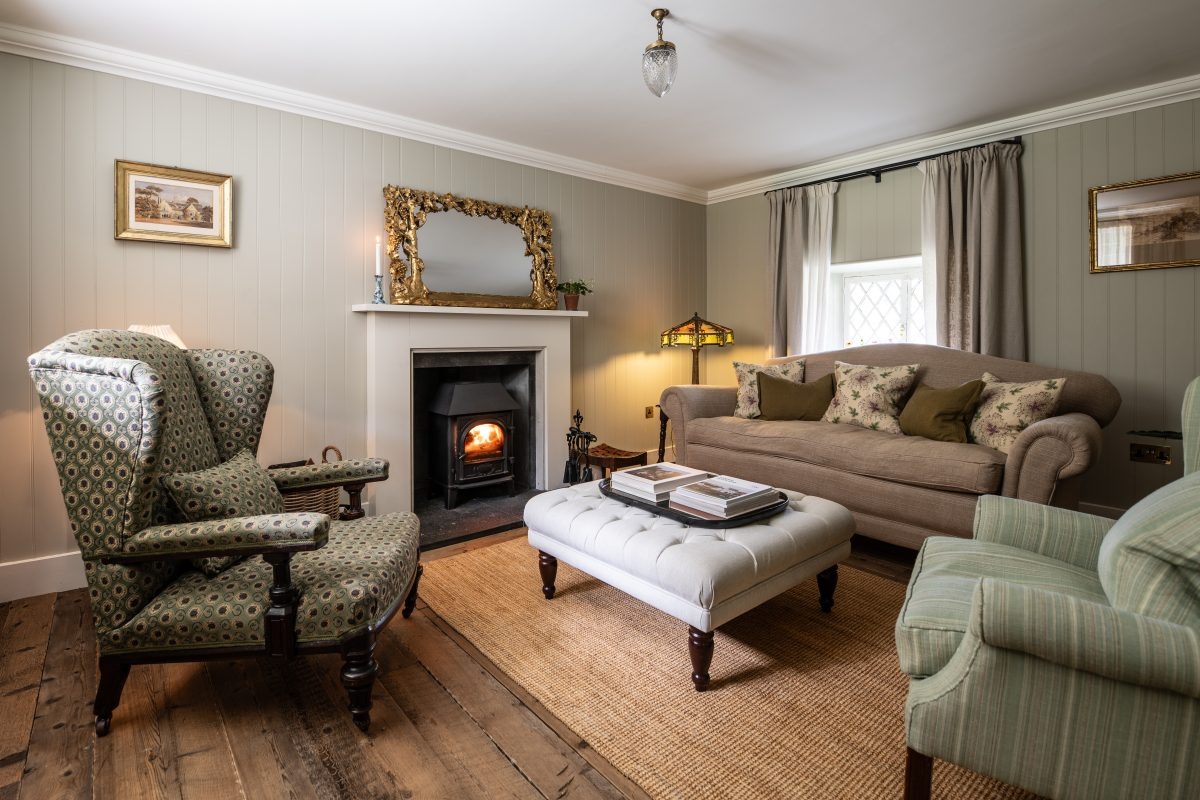 A green checked wing-back armchair and matching sofa flank a wood burning stove beneath a gilt mirror, a tufted footstool with books in the centre.