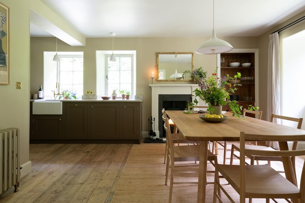 A wooden dining table set with flowers stands before an open-plan kitchen, a mantelpiece with a mirror and lit candle to the left.