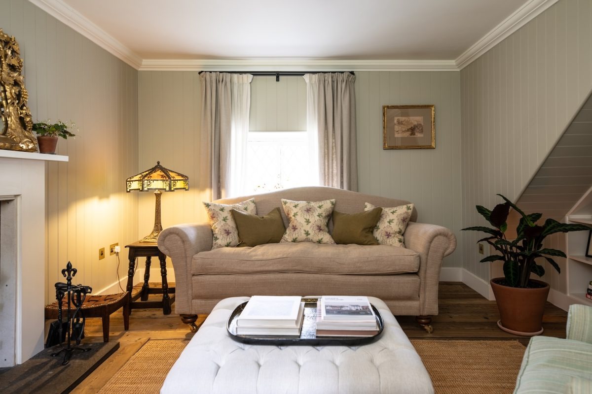 A beige sofa with floral cushions sits before a curtained window, a tufted footstool with books in front and a lit stained glass lamp beside the fireplace.