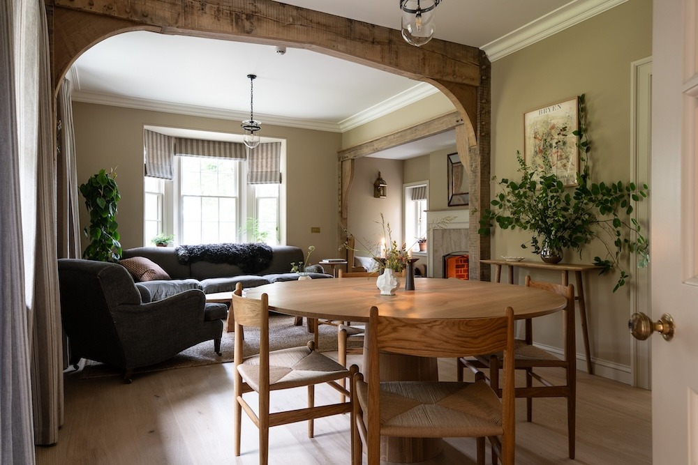 A round wooden dining table with woven-seat chairs stands beneath a beamed archway, a fireplace and grey sofa visible in the room beyond.