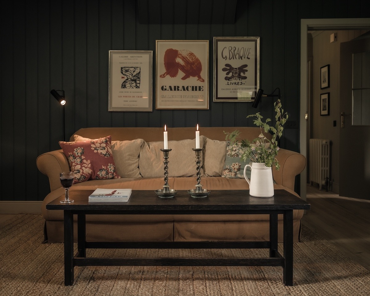 Framed prints including "Garache" hang above a tan sofa with floral cushions, lit candles and a jug of foliage on the table in front.