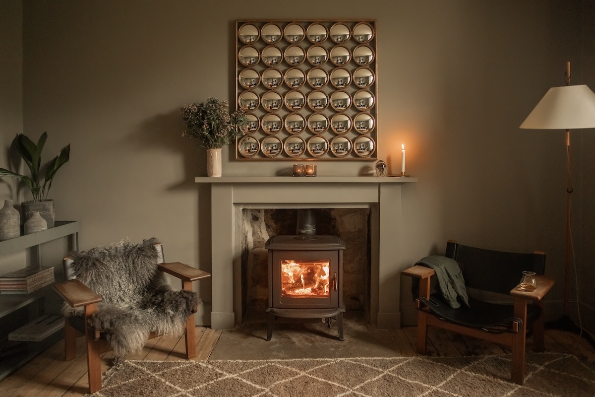 A wood burning stove burns beneath a framed grid of small circular botanical prints, a sheepskin-draped chair and a lit candle to the sides.