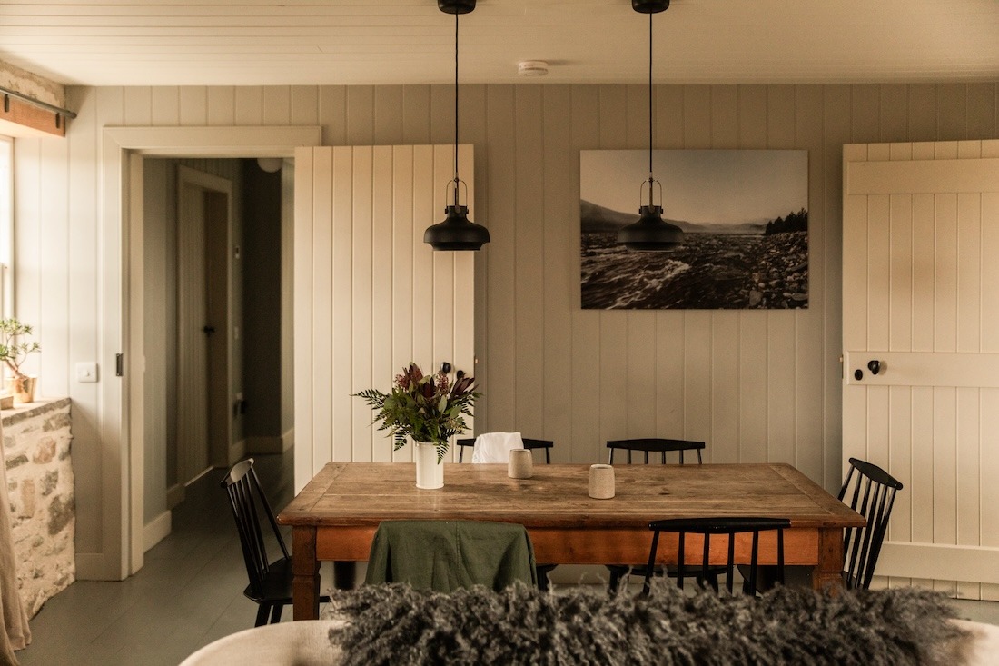A wooden dining table set with flowers and candles stands before panelled walls hung with a framed landscape photograph, black pendant lights above.