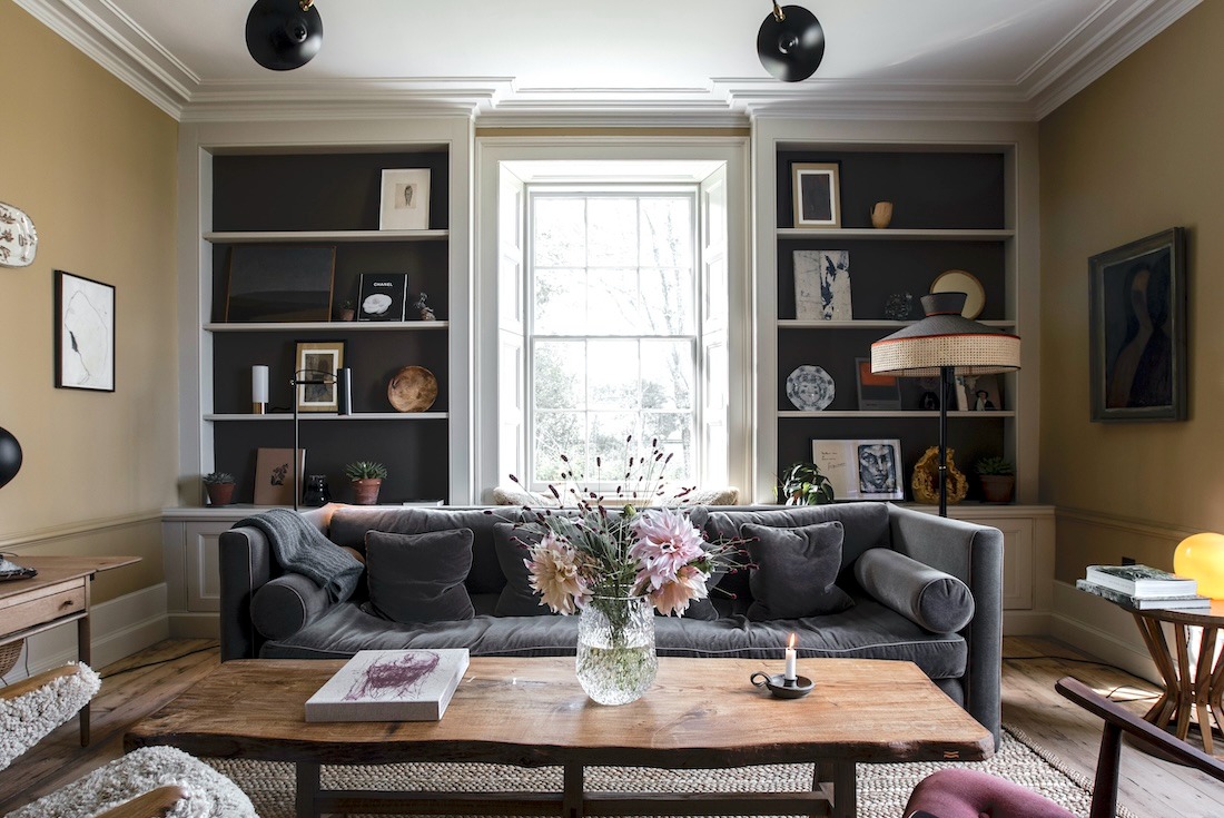 A dark grey sofa sits below built-in bookshelves and a curtained window, a vase of pink flowers and lit candle on the wooden coffee table.