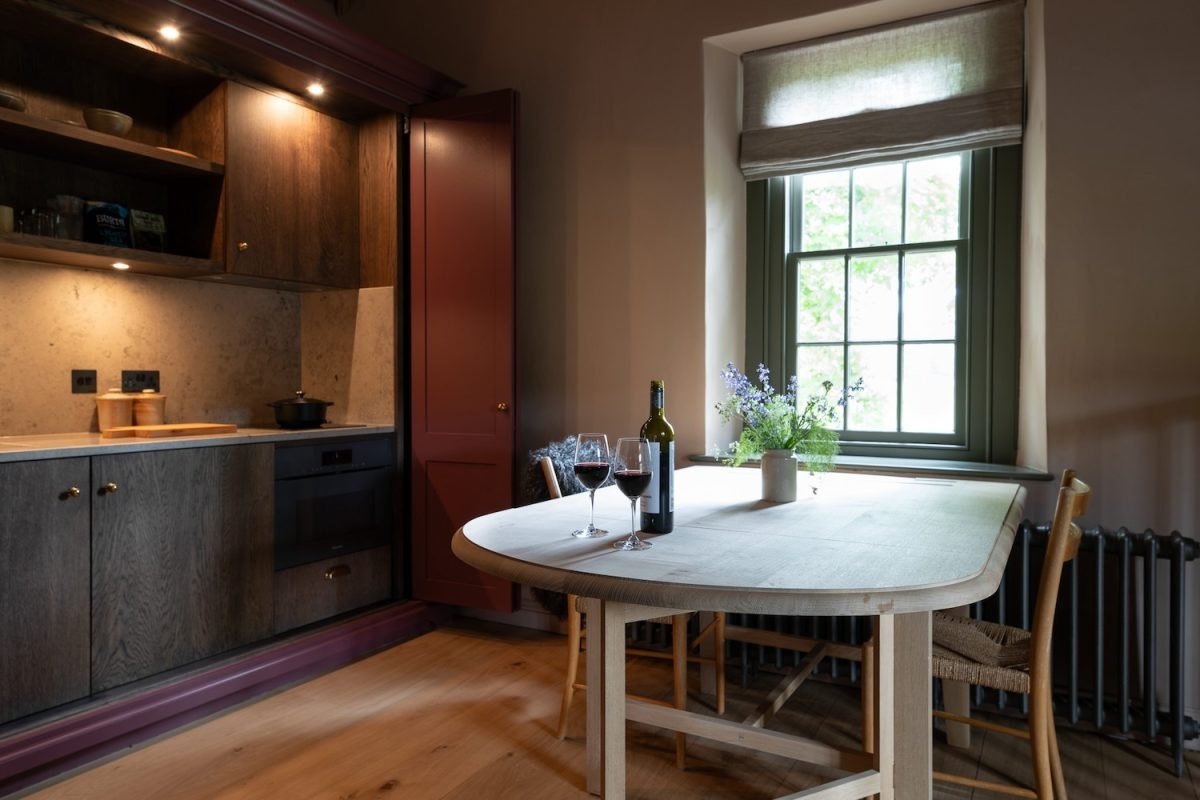 A round wooden table set with wine and flowers stands before a window with a pleated blind, a dark wood kitchen to the left.