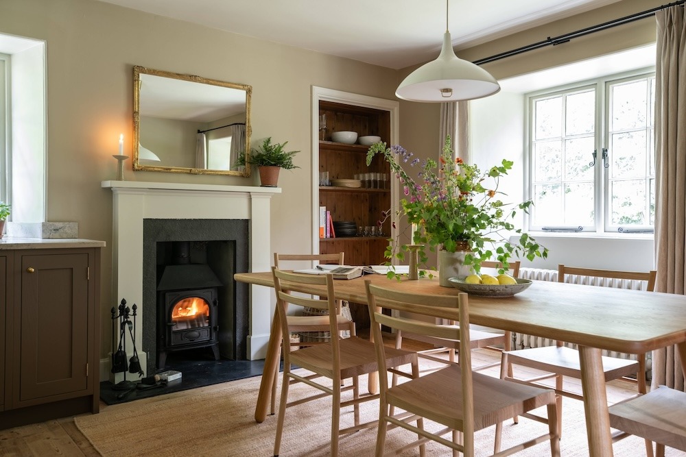 A wood burning stove is alight below a gilt-framed mirror in a dining room, a table set with flowers and a dresser of crockery to the side.
