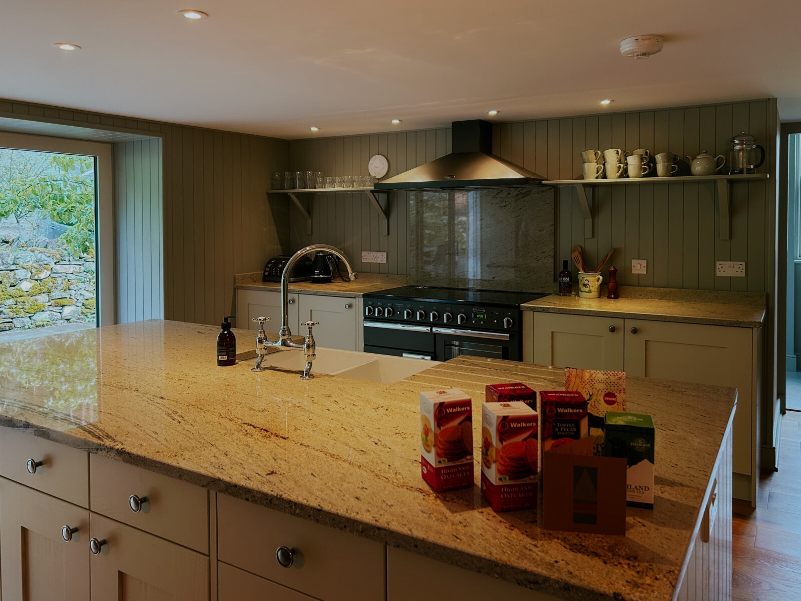 A kitchen island with a marble worktop stands before a range cooker and extractor hood, open shelving of mugs and glasses on the green panelled wall.