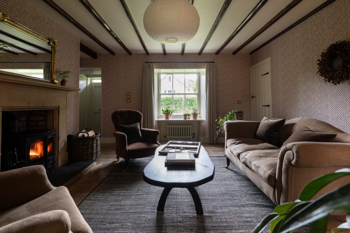 Sofas and an armchair surround a low table before a lit fireplace, beamed ceiling and patterned wallpaper completing the sitting room.