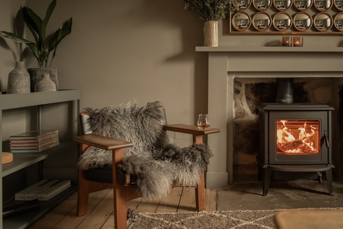 A sheepskin-draped armchair sits beside a wood burning stove set in a green fireplace, a shelf of ornaments above and a plant on a console table to the left.