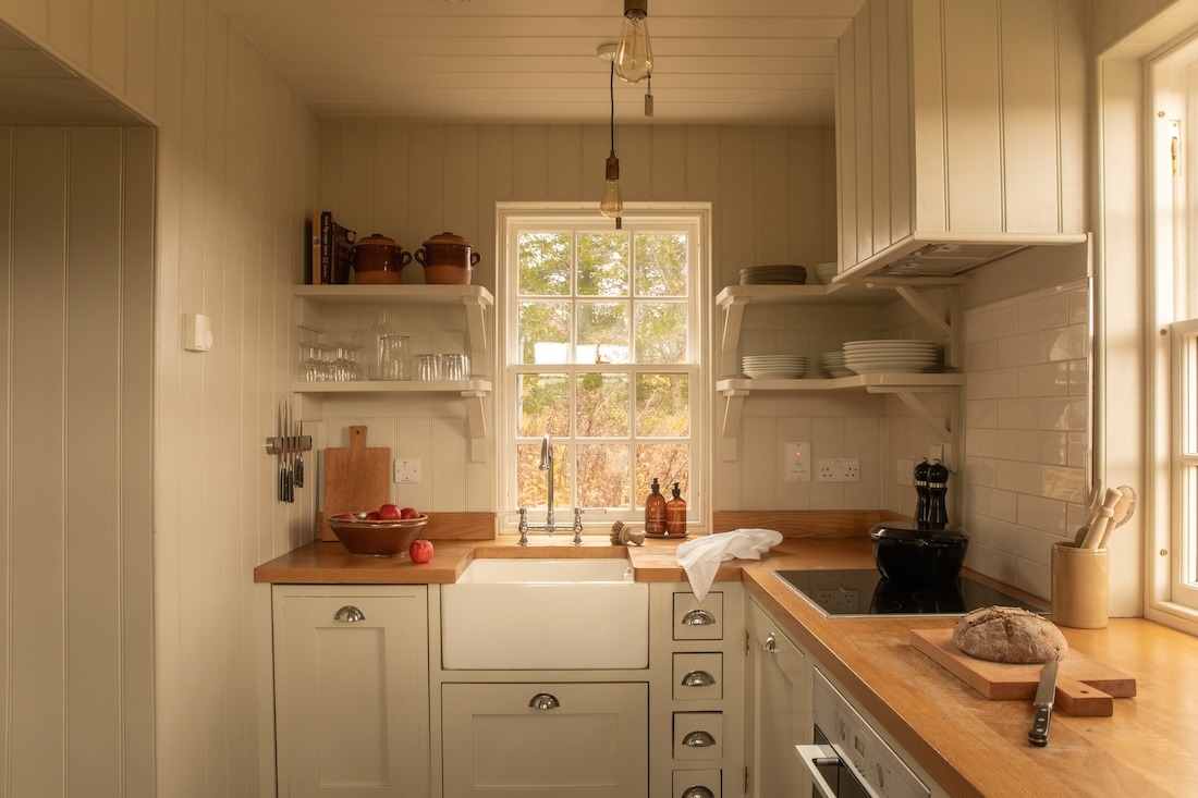 A galley kitchen with panelled cabinetry and open shelving of crockery leads to a window, a chopping board of apples on the wooden counter.