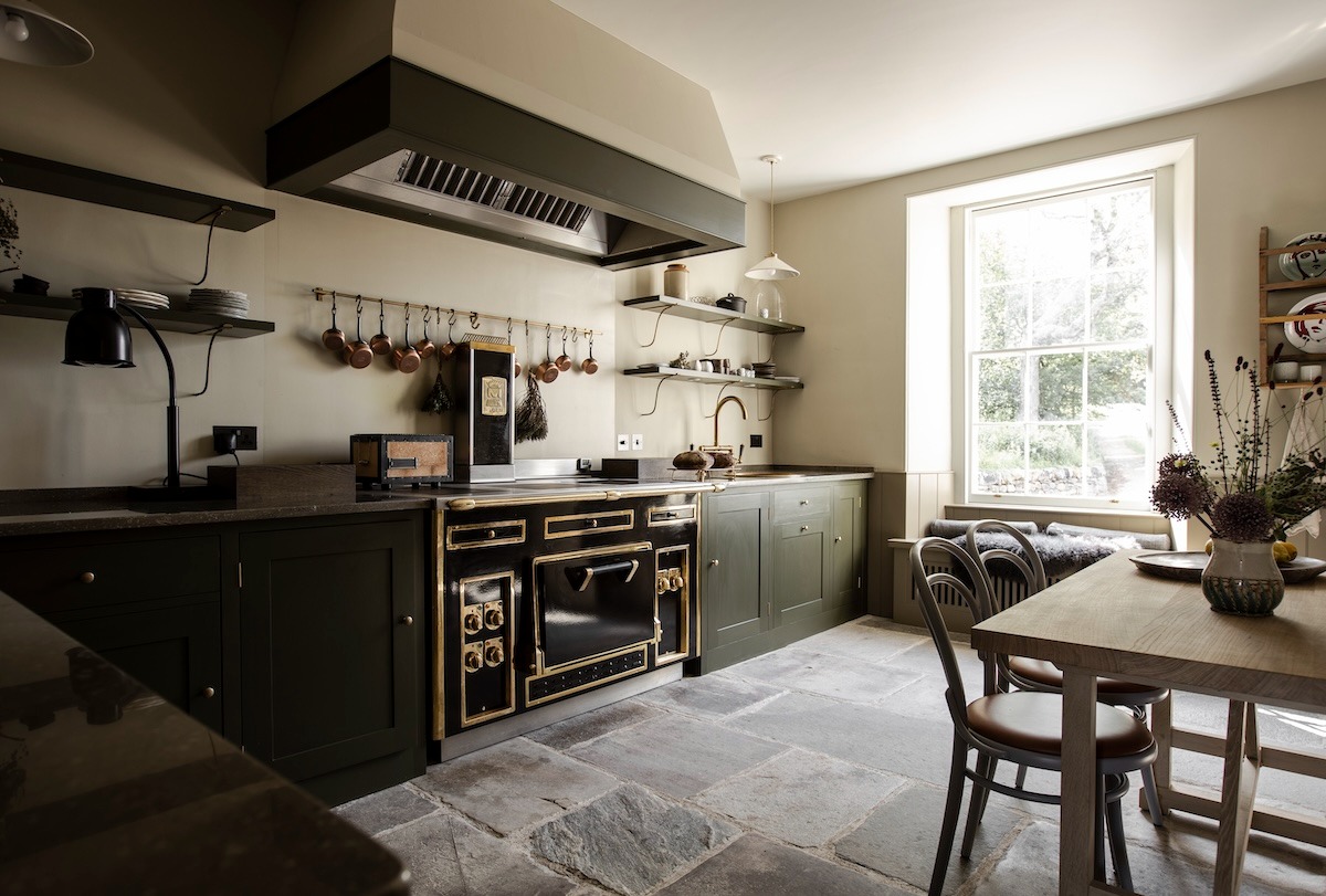 A black range cooker with brass fittings stands below a matching extractor hood, copper pans hanging above and a dining table to the right.